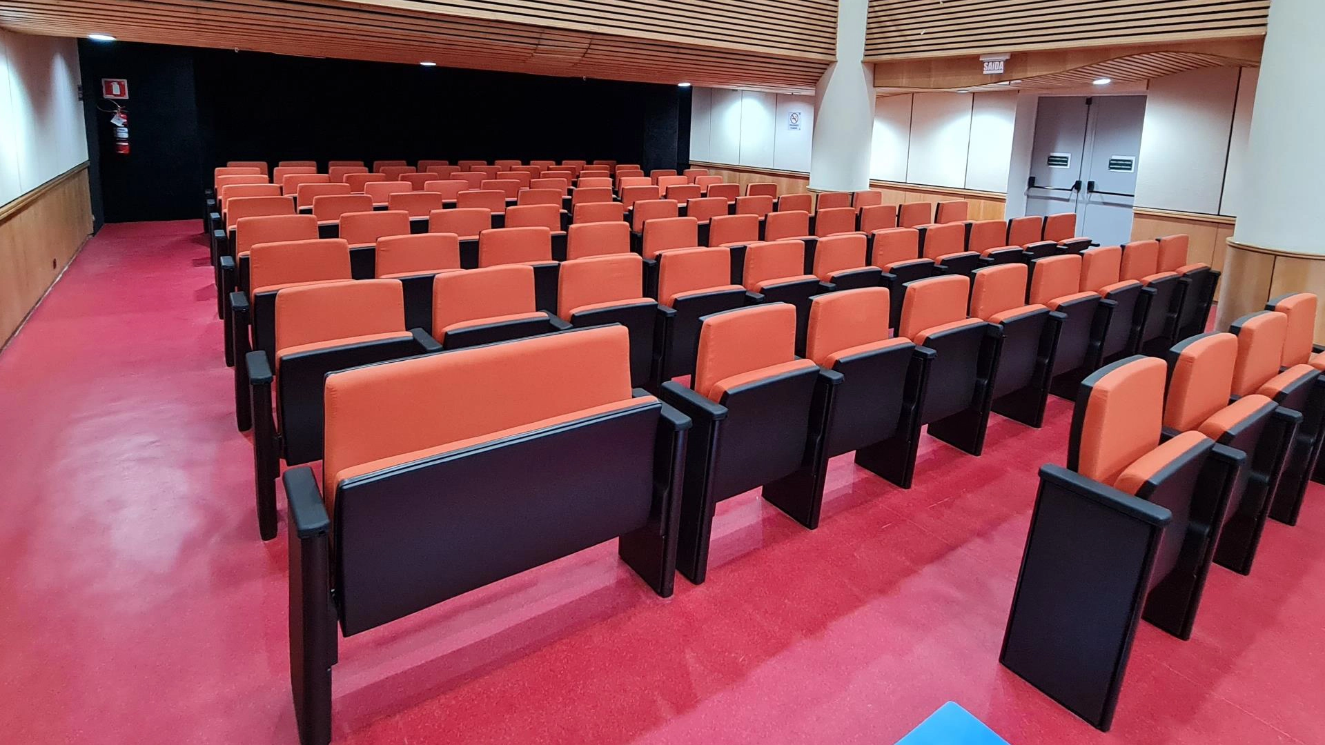 Corredor lateral de auditório com cadeiras estofadas e piso vermelho
